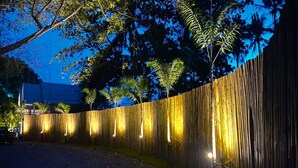 A wooden fence with vertical slats and embedded lights, a curved driveway, and trees in the background. - Slumber Party Beach Koh Phangan (Ko Pha-ngan)
