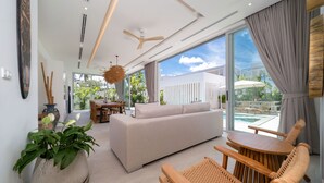 A modern living room with a sofa, wooden chairs, a dining table, and a large window with a view of the outdoors. - Modern Open Design 4BR Pool Villa Mina (Rawai)