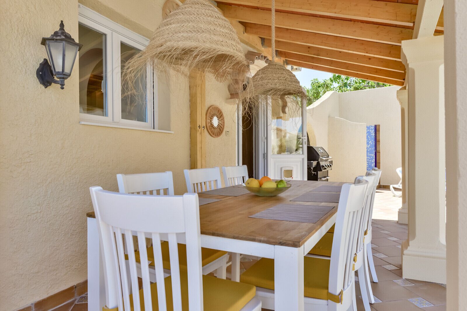 A wooden table with white chairs, a bowl of fruit, and a grill outside.