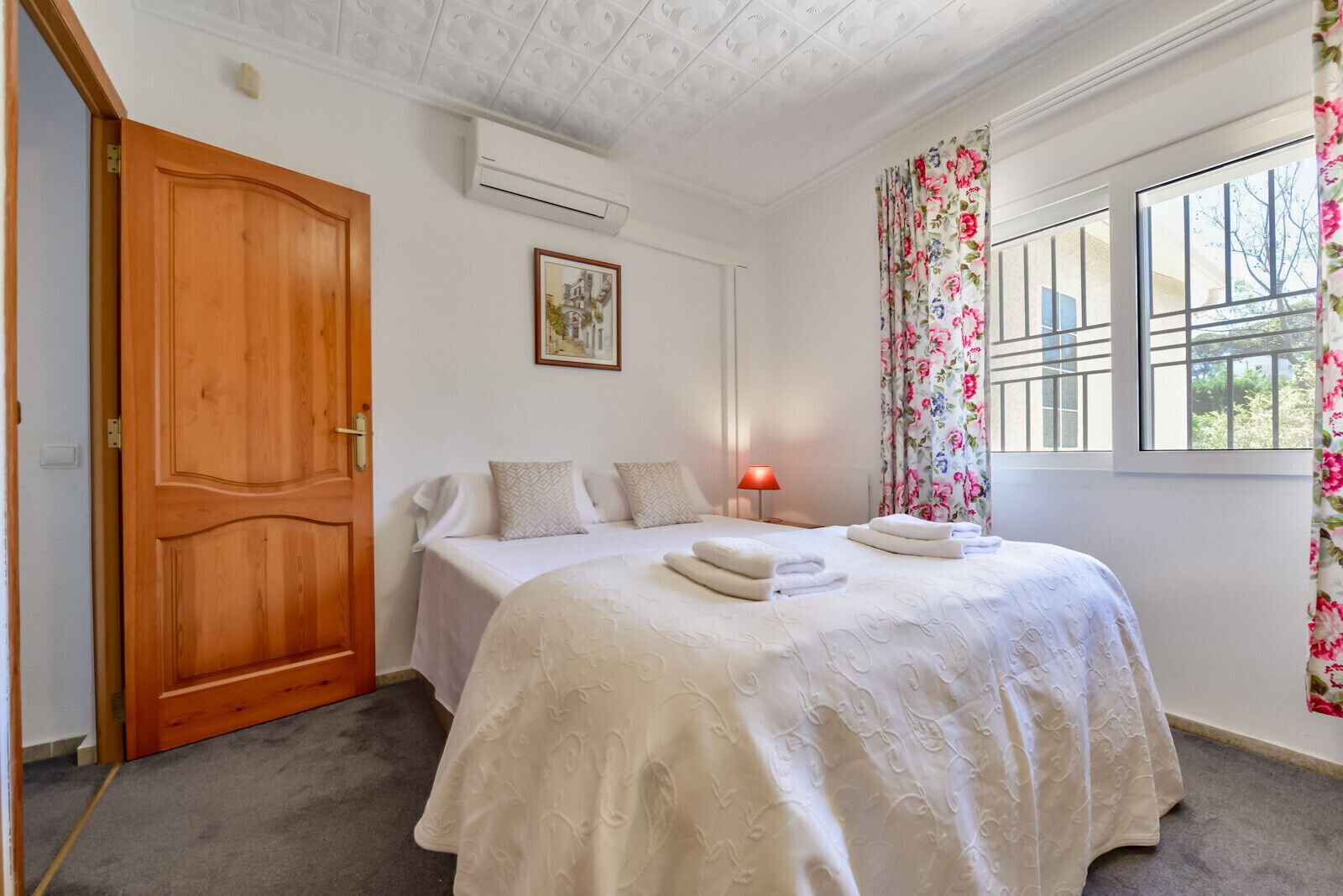 A bedroom with a wooden door, a bed with white linens, a framed picture on the wall, a window with floral curtains, and an air conditioning unit.