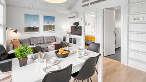 Een moderne eetruimte met een witte tafel, zwarte stoelen en een hangende pendantlamp. Er is een grijze bank en een kleine plant op de tafel.