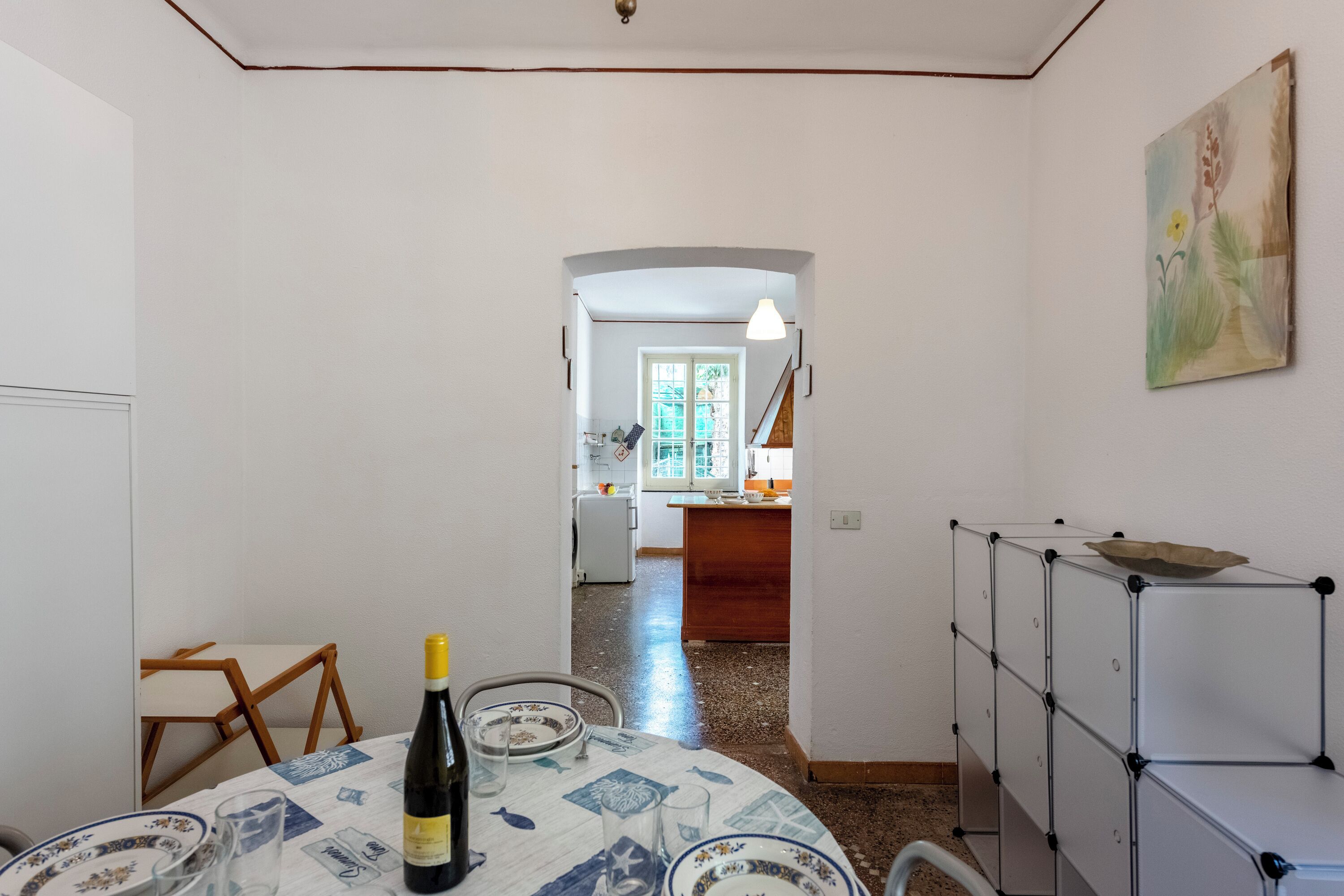 A dining area with a table set for two, a bottle of wine, and a painting on the wall.