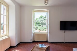 Egy nagy ablakos, radiátorral felszerelt szoba, melyben a falra van szerelve egy síkképernyős televízió, és egy fonott szék található.