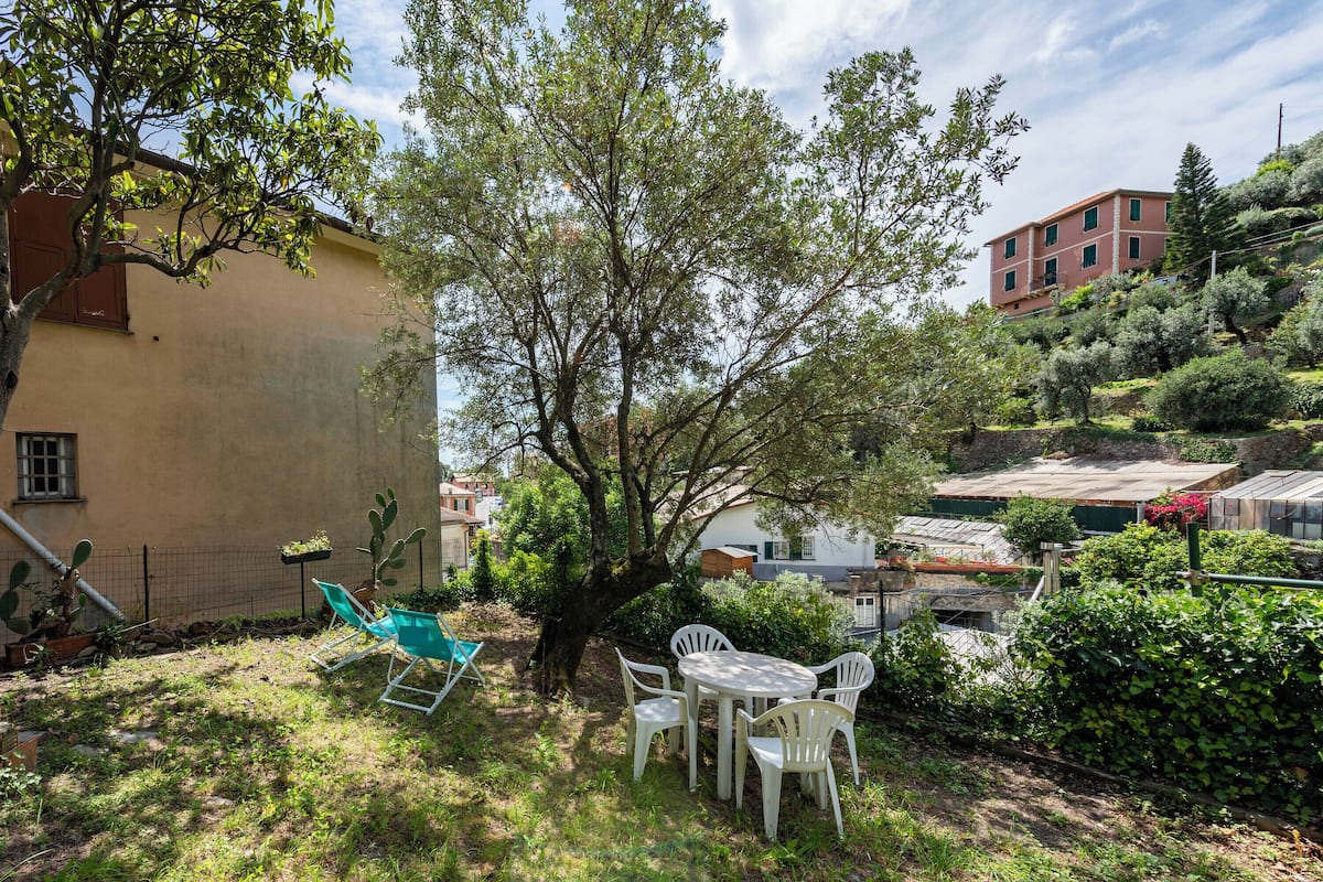 A patio with a table and chairs, surrounded by greenery and a building in the background.