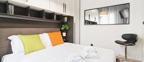 A modern hotel room with a large bed, a bedside table with a lamp, a mirror, and a black chair.