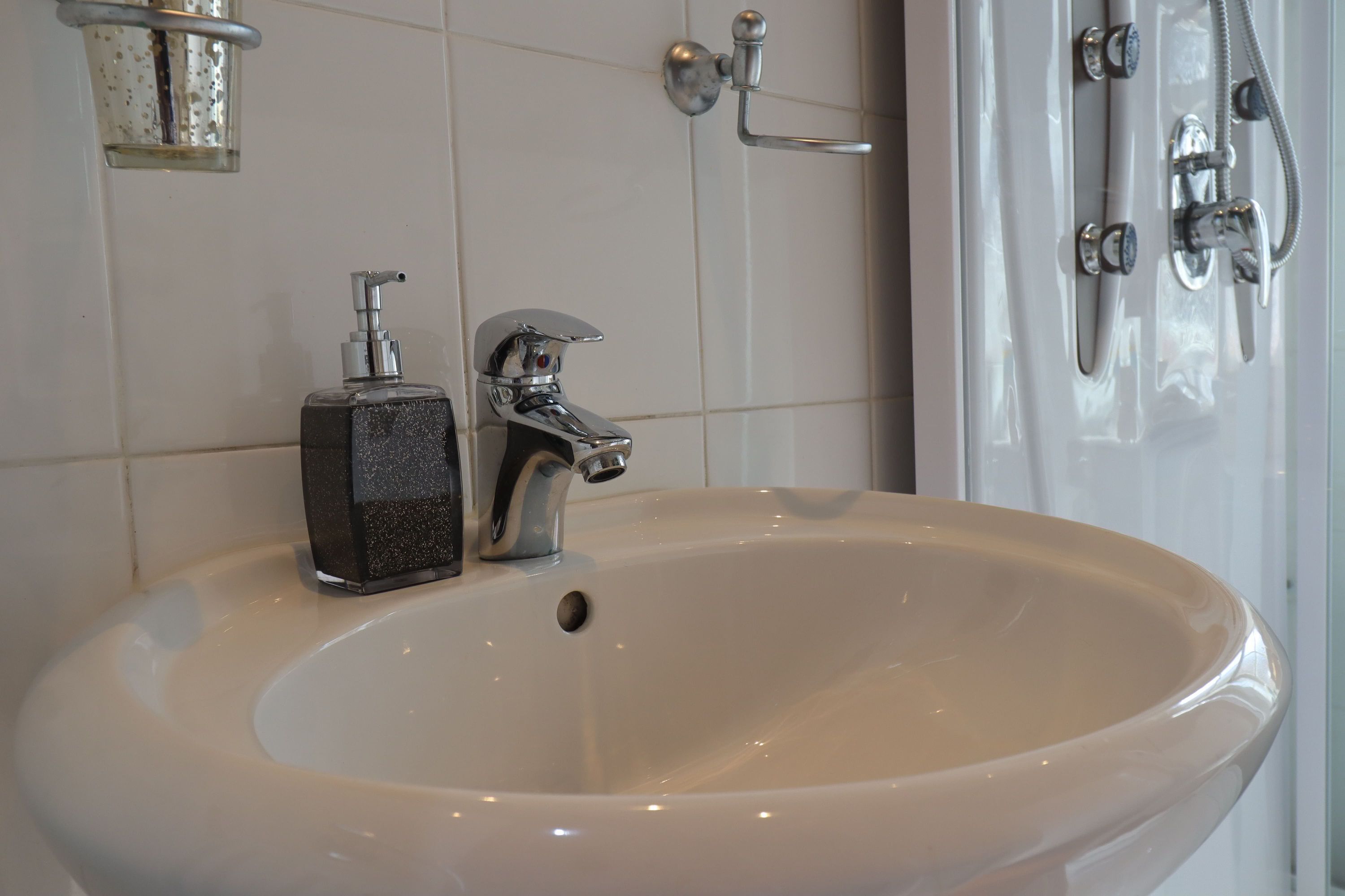 Une salle de bain avec un lavabo blanc, un distributeur de savon et une douche équipée d’un pommeau de douche à main.