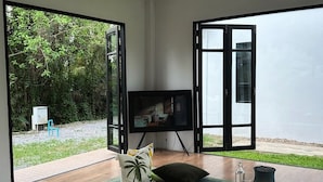 A spacious room with a large sliding glass door leading to a garden, a flat-screen TV, and a minimalist coffee table. - Waste Land Homestay & Resort Chiangmai (San Pa Thong)