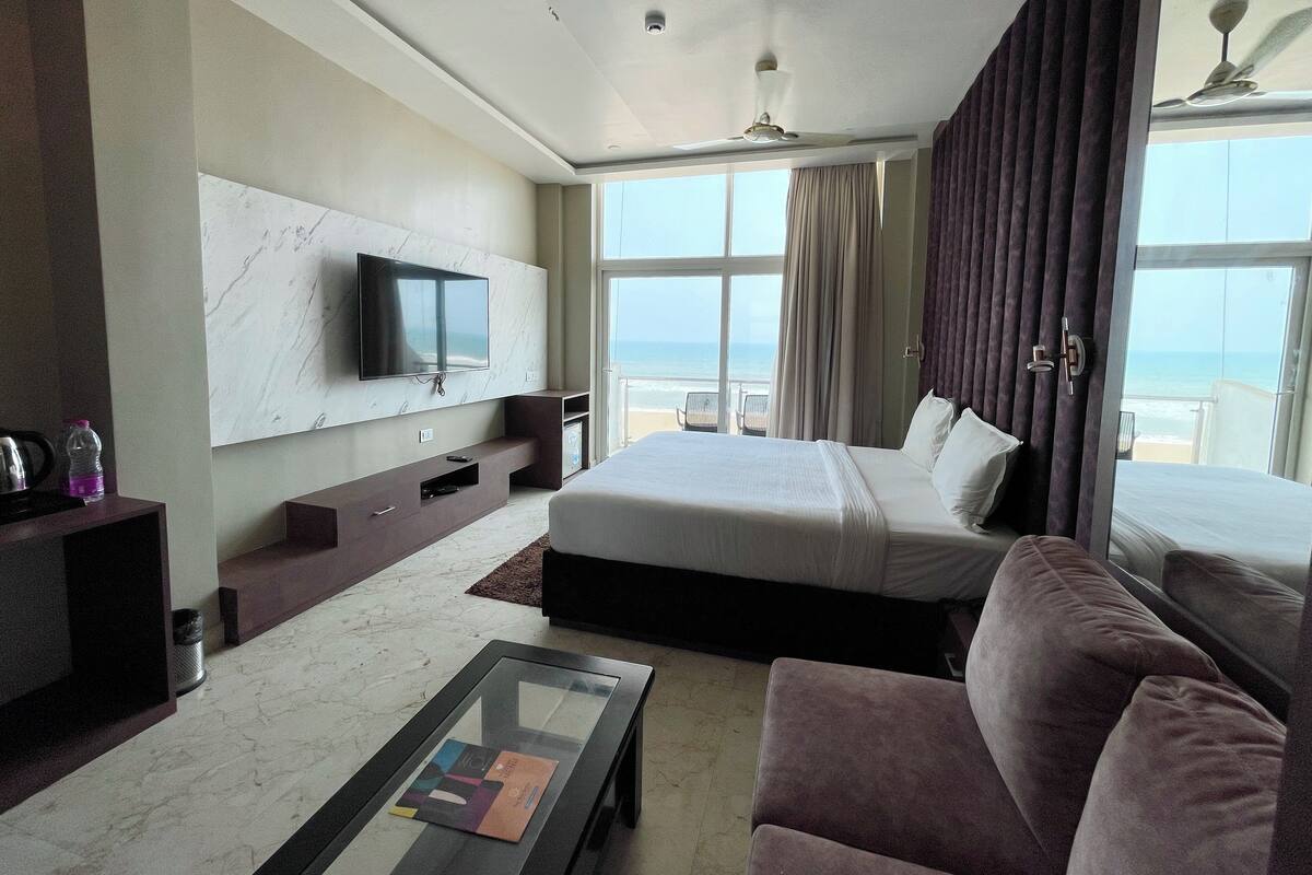 Quarto de hotel com uma cama grande, televisão fixada na parede, mesa de centro de vidro e vista para o oceano.