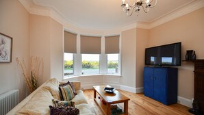 A living room with a sofa, a coffee table, a TV, and a bay window with curtains. - Arran View in Prestwick (Prestwick)