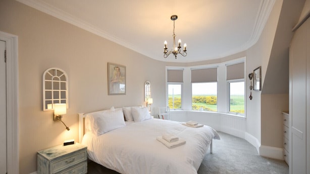 A bedroom with a large bed, a nightstand, a chandelier, and a view of the outdoors through large windows. - Arran View in Prestwick (Prestwick)