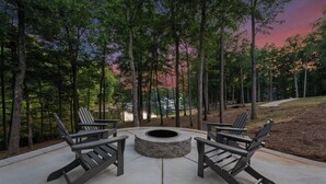 A circular stone fire pit with four Adirondack chairs around it, set on a concrete patio surrounded by trees. - Lakefront Retreat with Private Dock, Pool, Game Room | Sleeps 18 (Mooresville)