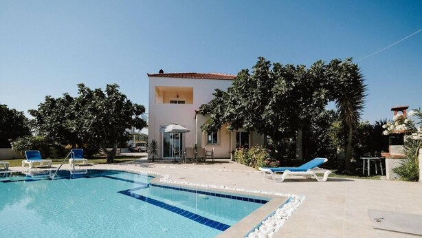 A swimming pool with lounge chairs, a building with a tiled roof, and surrounding greenery. - Villa Maia in Rhodes (Rhodes)
