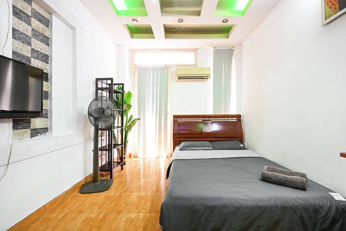 Un dormitorio con cama, televisor, ventilador y ventana.