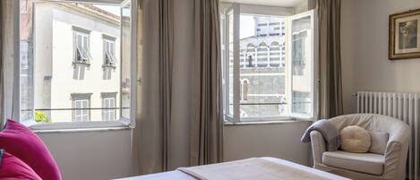 A bedroom with a bed, a window with curtains, a chair, and a radiator.