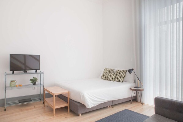Une chambre à coucher au style minimaliste, avec un lit, un téléviseur posé sur une étagère, une petite table et une lampe.