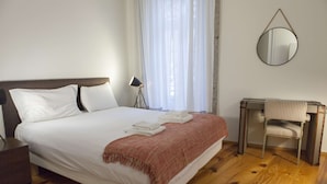 Una cama bien hecha con sábanas blancas y una manta rosa, una mesita de noche con lámpara, una silla y un espejo en la pared.