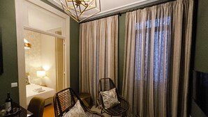 Habitación de hotel con una cama, dos sillones de mimbre con cojines, una mesita redonda, una lámpara y vistas al exterior a través de cortinas.