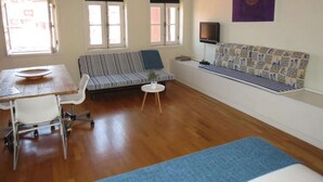 Une pièce avec un plancher en bois, un canapé à rayures, une petite table ronde, une chaise blanche et un téléviseur fixé au mur.