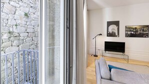 A modern living room with a stone wall, a glass door leading to a balcony, a grey sofa, a TV on a stand, and framed pictures on the wall. - Suite with a double bed and air conditioning (Porto)