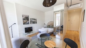 A modern living room with a sofa, a glass coffee table, and a TV. There are two chairs, a bed with a headboard, and a window with curtains. - Suite with a view to the city, double bed and air conditioning (Porto)