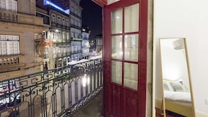 Un balcón con una puerta roja, una cama con cabecera blanca y vistas a una calle iluminada de noche.