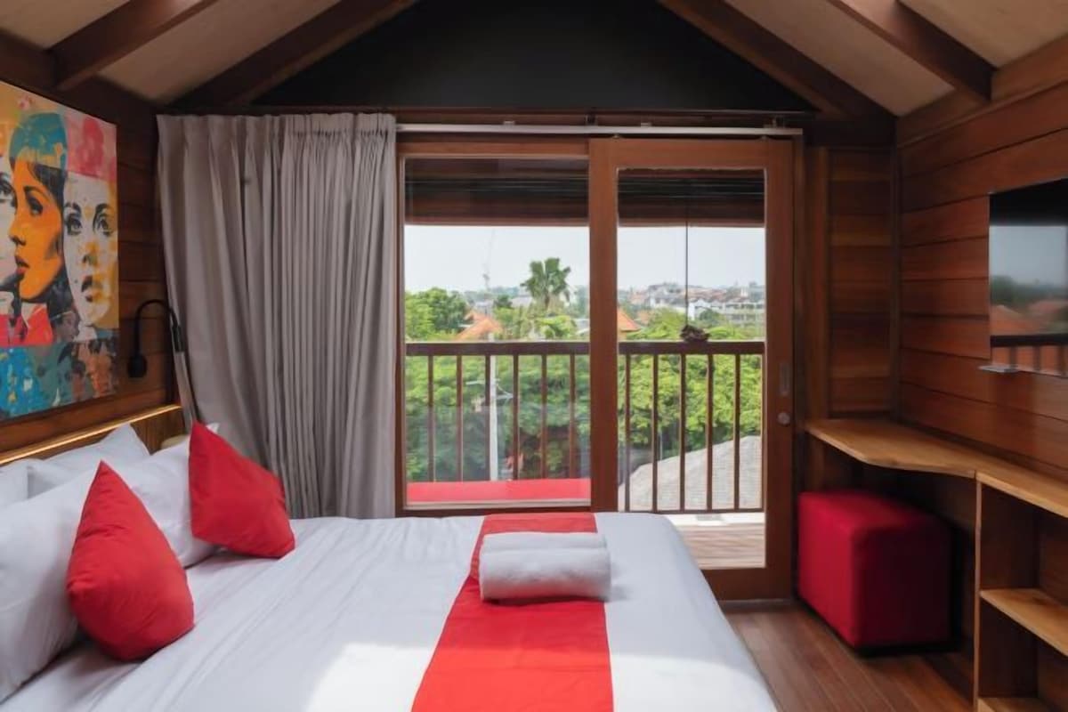 A hotel room with a bed, a red and white striped bedspread, a red pillow, a white pillow, a red ottoman, a wooden TV stand, and a painting on the wall.