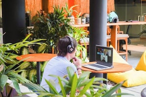 A person using a laptop with a video call on a round table in a room with plants and wooden furniture. - SOKKOOL CANGGU - COWORKING & COLIVING (Canggu)