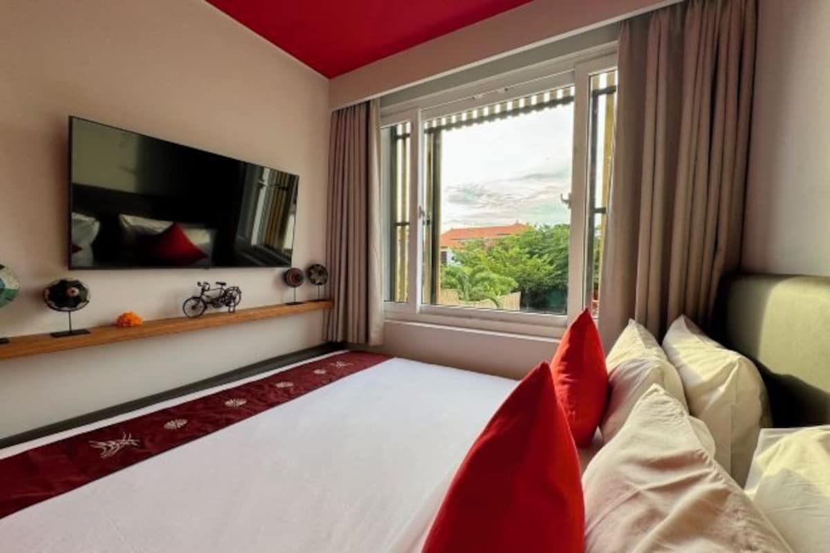 A hotel room with a bed, a television mounted on the wall, a window with curtains, and a view of greenery outside.