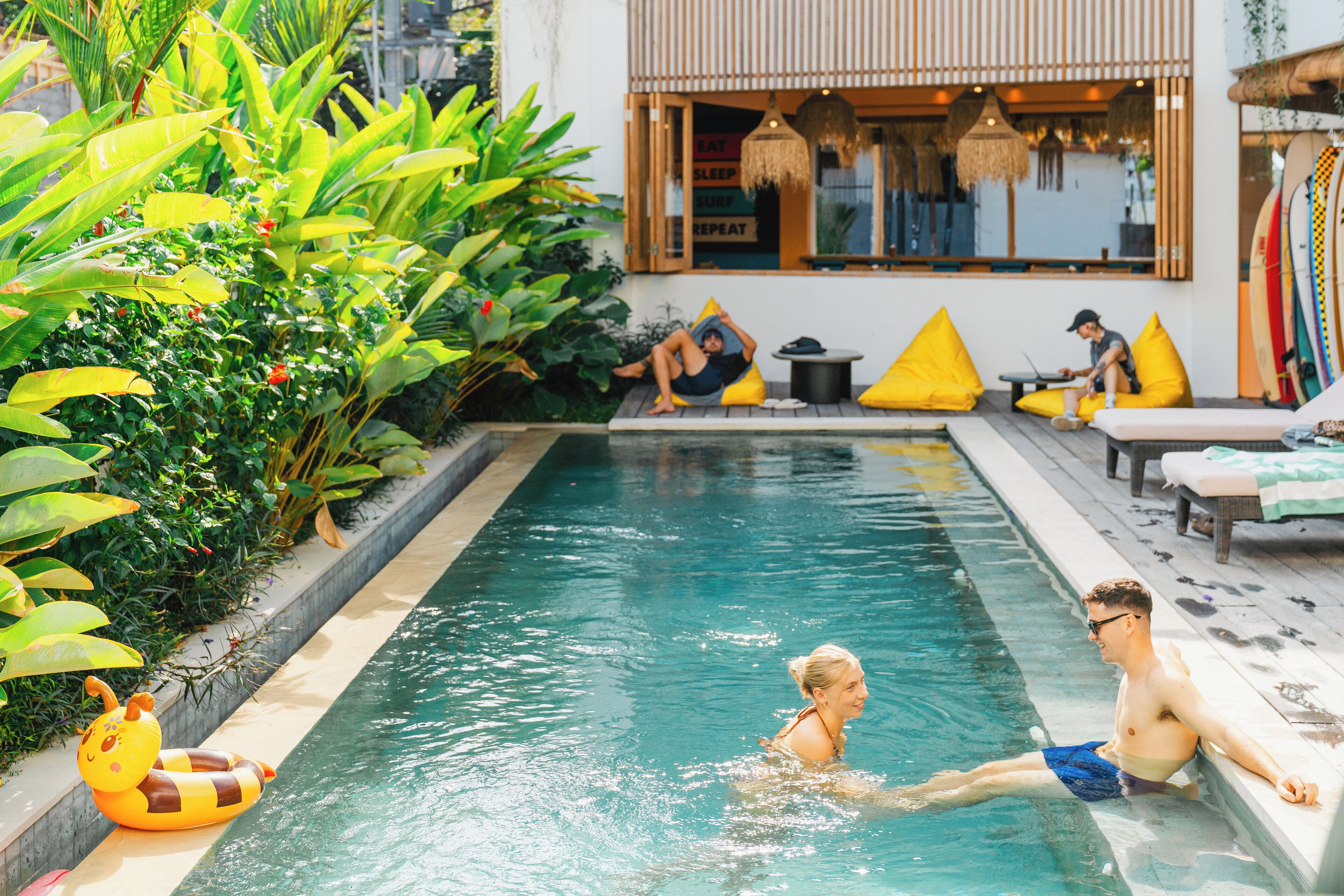 A pool area with a lounge chair, a small table, and a person sitting by the pool.