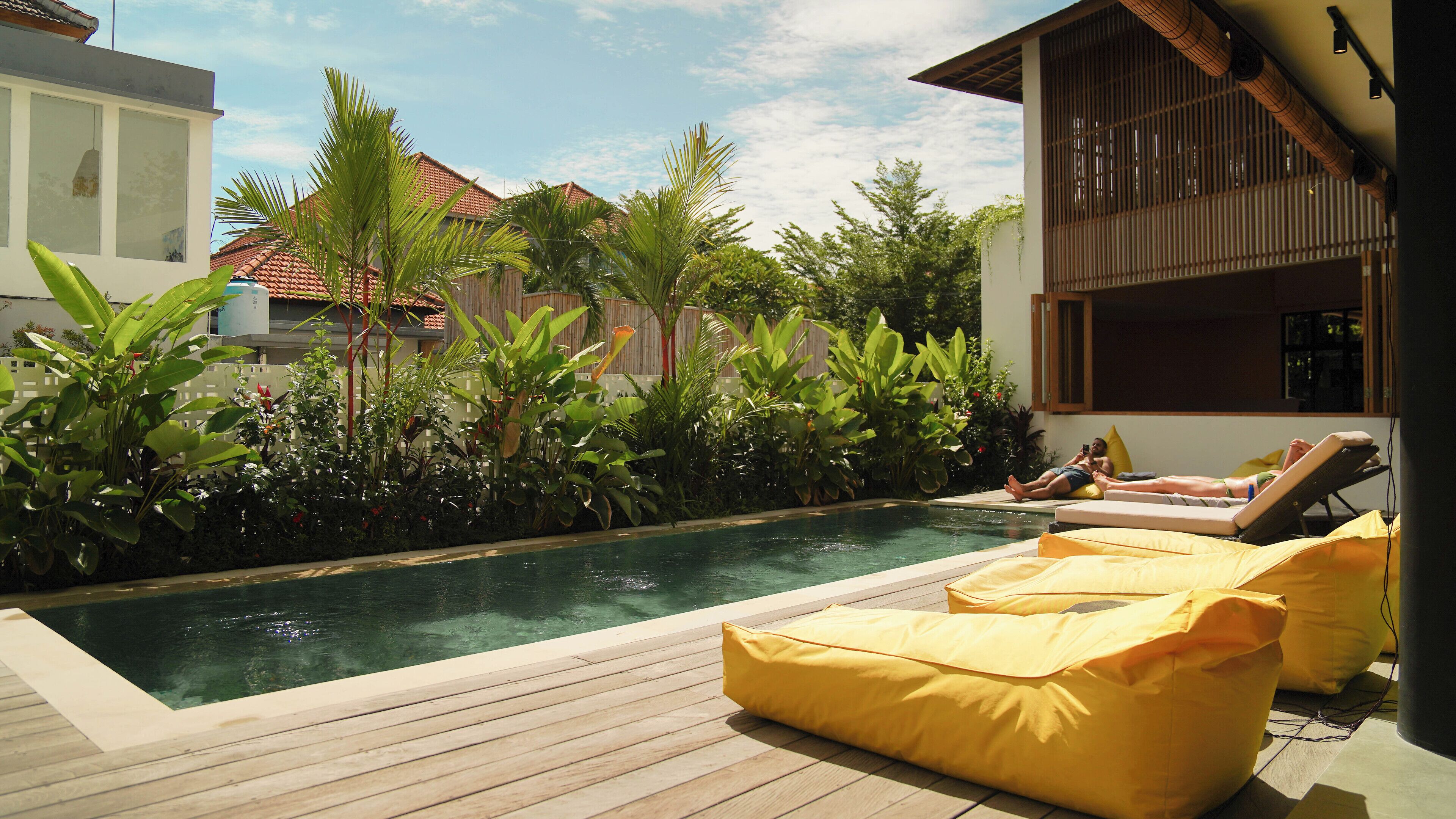 A pool area with wooden deck, yellow cushions, and a view of a building with a tiled roof.