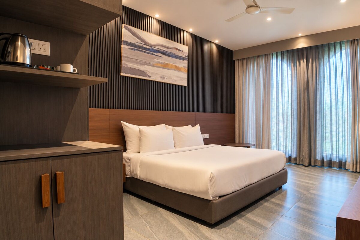 A modern hotel room with a large bed, a flat-screen TV, a desk with a lamp, and a view of the outdoors through the window.