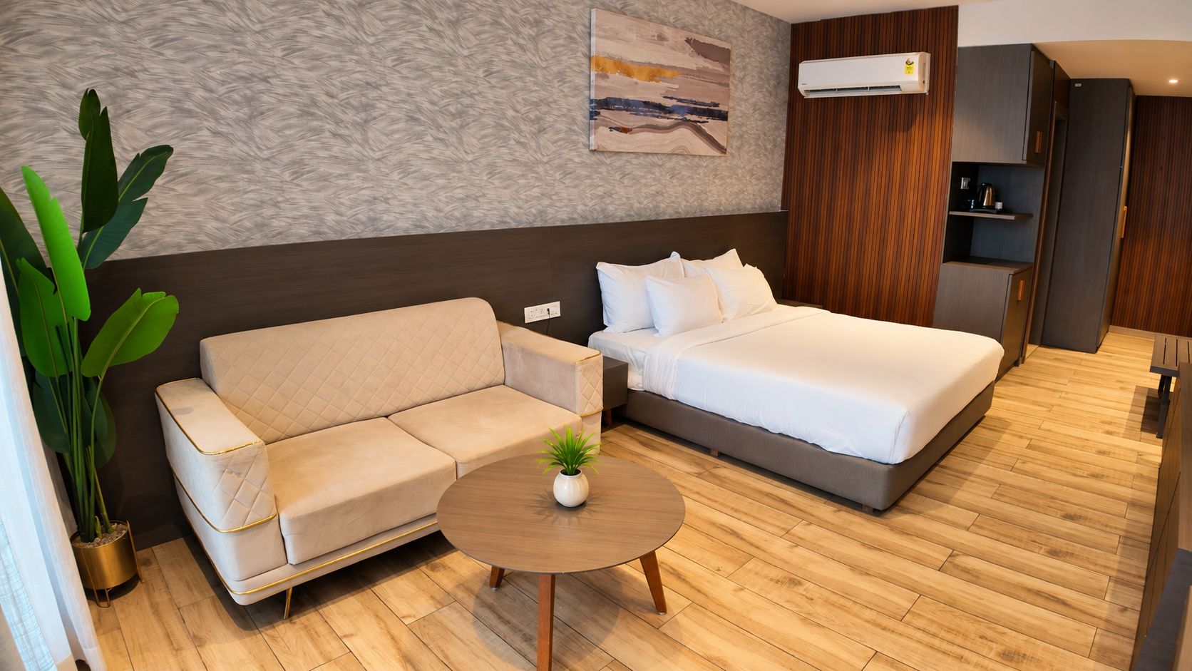 A modern hotel room with a sofa, a bed, a small round table with a plant, and a wooden floor.