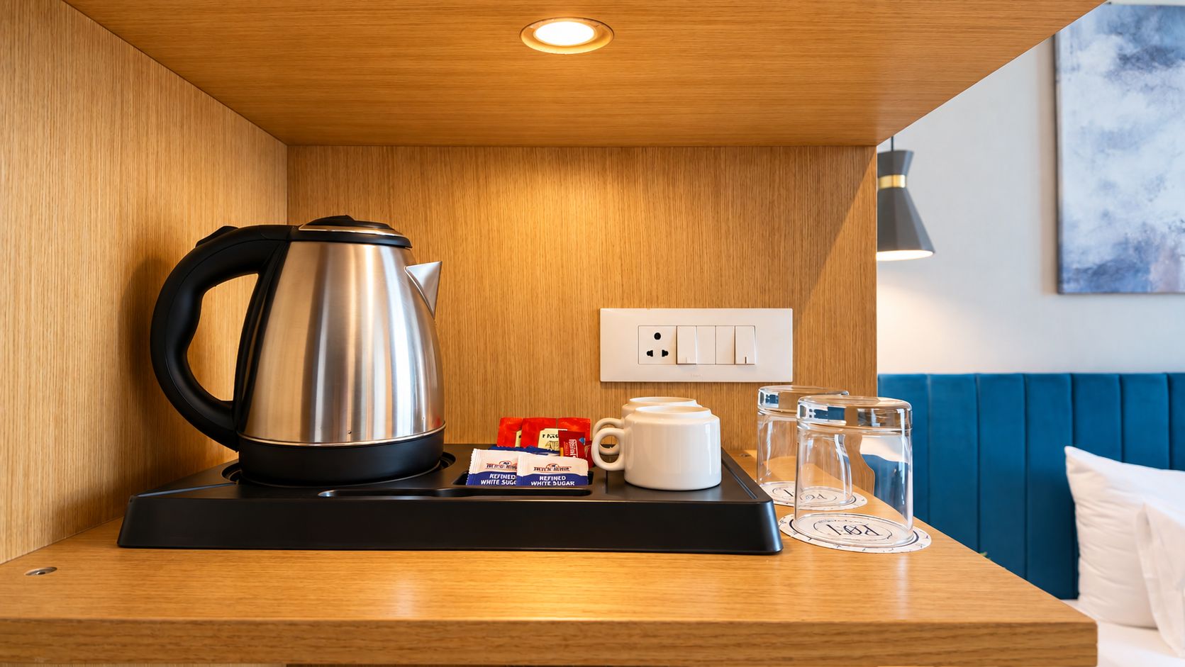 A hotel room with a wooden countertop, a kettle, sugar packets, a mug, and a glass pitcher.