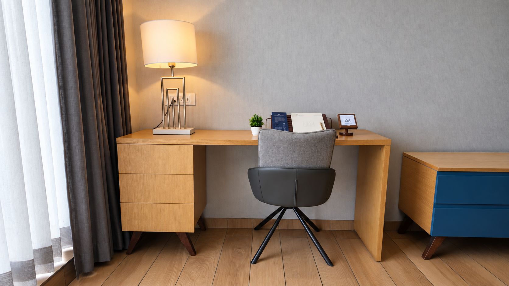 Ein Hotelzimmer mit einem Holztisch, einem Stuhl, einer Lampe, einer kleinen Pflanze und einer Uhr.