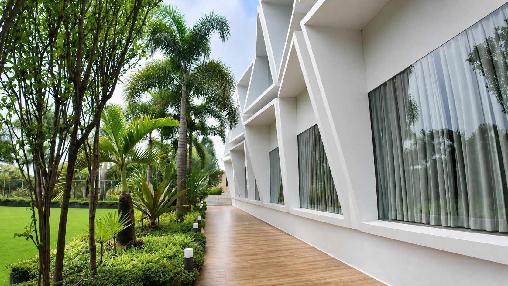 A modern hotel corridor with large windows, a wooden floor, and a well-maintained garden with palm trees.