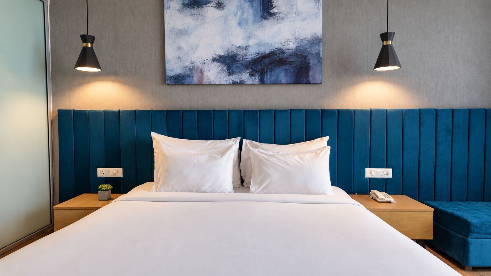 A hotel room with a large bed, two bedside tables, a blue headboard, a painting on the wall, and two pendant lights.