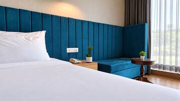 A hotel room with a large bed, a blue upholstered headboard, a bedside table with a phone and a plant, and a small round table with a plant.