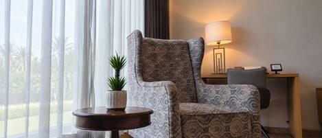 A room with a patterned armchair, a small round table with a plant, a lamp, and a desk with a chair.