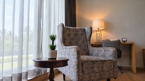 A room with a patterned armchair, a small round table with a plant, a lamp, and a desk with a chair. - THE DEMOCRACY HOTEL & RESORT (Nashik)