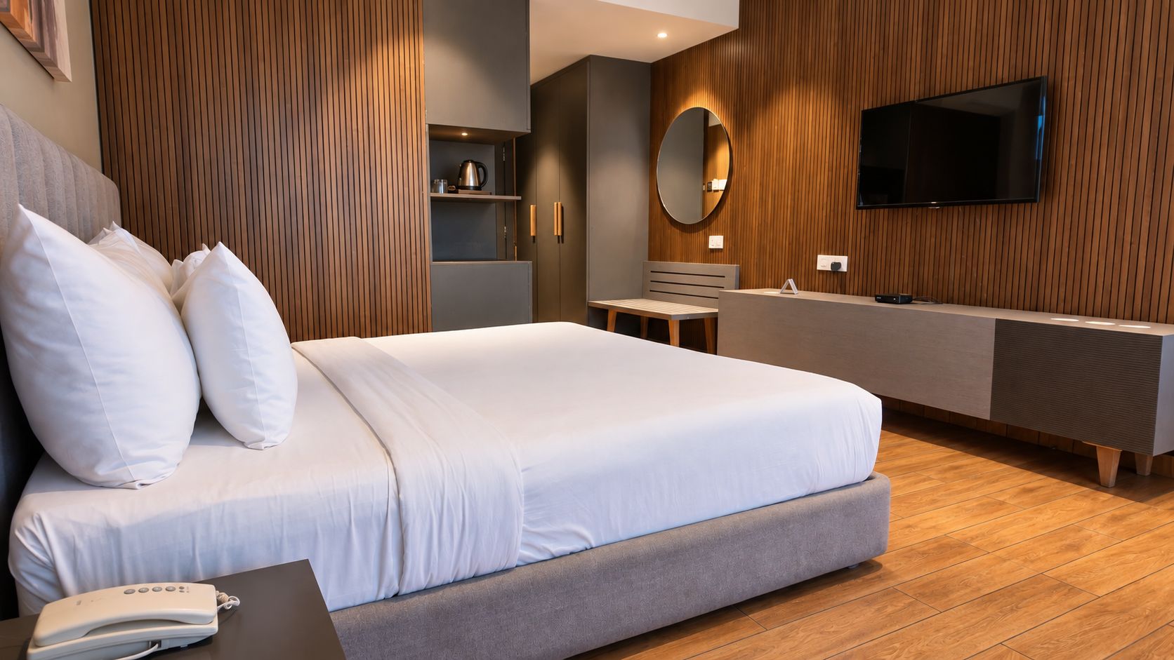 A modern bedroom with a large bed, a bedside table, a TV mounted on the wall, and a wooden floor.