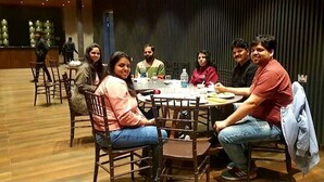 A group of people seated around a round table in a room with wooden flooring and a dark acoustic panel wall. - THE DEMOCRACY HOTEL & RESORT (Nashik)