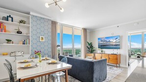 A modern living room with a dining area, large windows, a flat-screen TV, and a view of the outdoors.