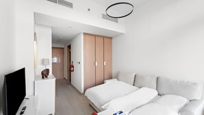 Moderni hotellihuone, jossa on taulutelevisio, pieni pöytä, jossa on hedelmiä ja rasia, sohva ja puinen vaatekaappi.