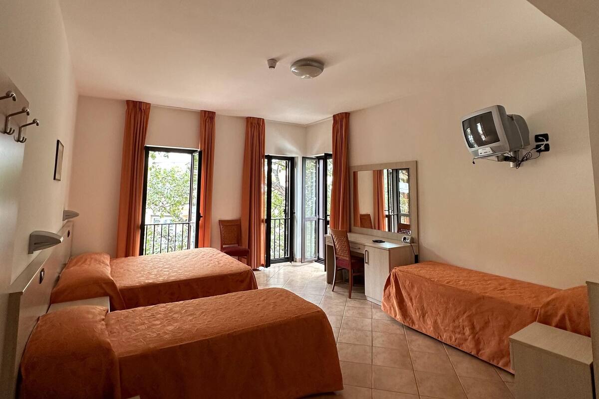 Hotellihuone, jossa on kaksi sänkyä, työpöytä, tuoli, televisio ja parveke, jossa on verhot.
