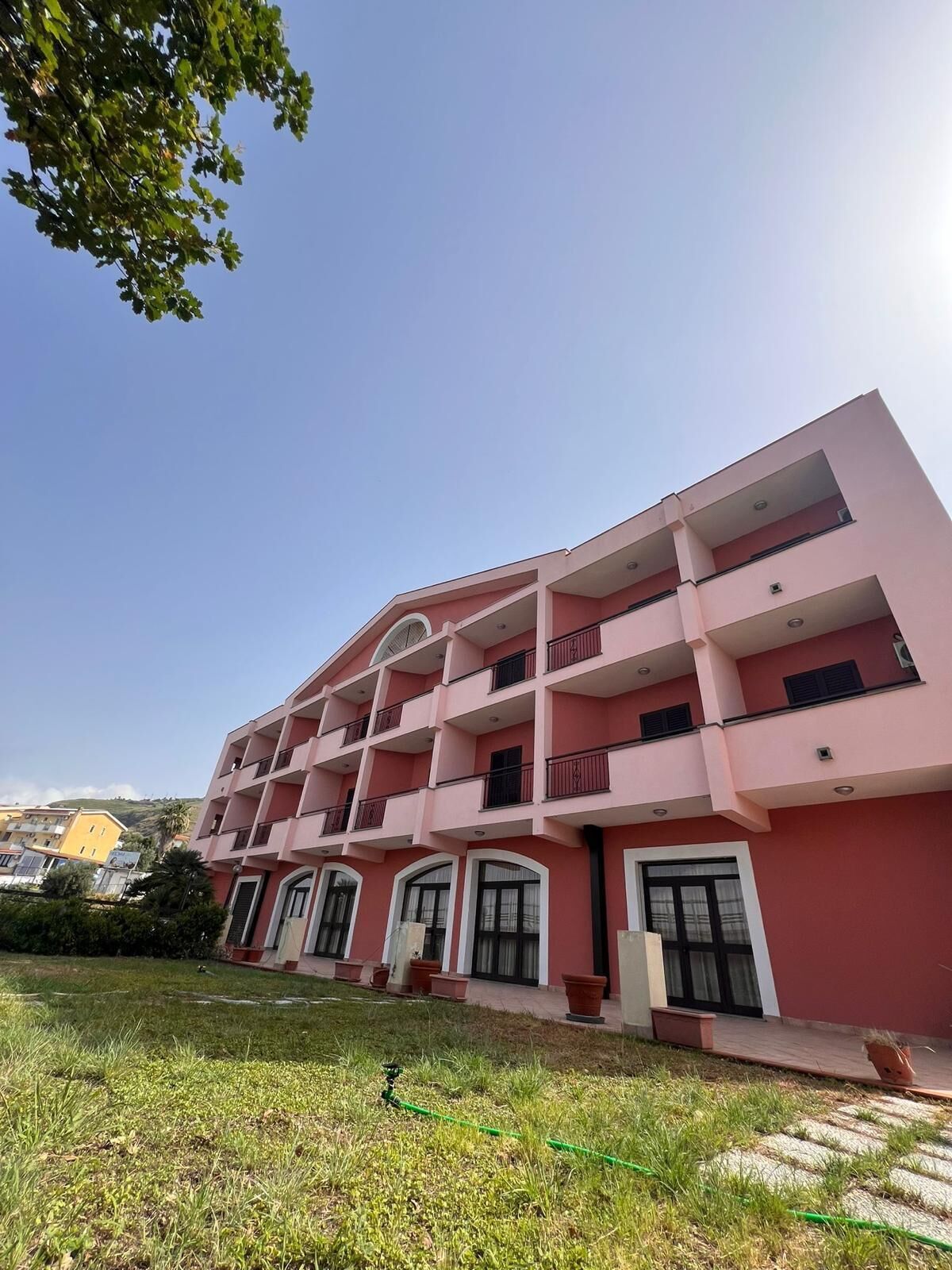 A multi-story pink building with balconies, large windows, and a grassy area in front.