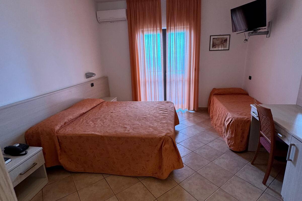 Hotellihuone, jossa on kaksi sänkyä, työpöytä, tuoli, televisio ja ikkuna, jossa on verhot.