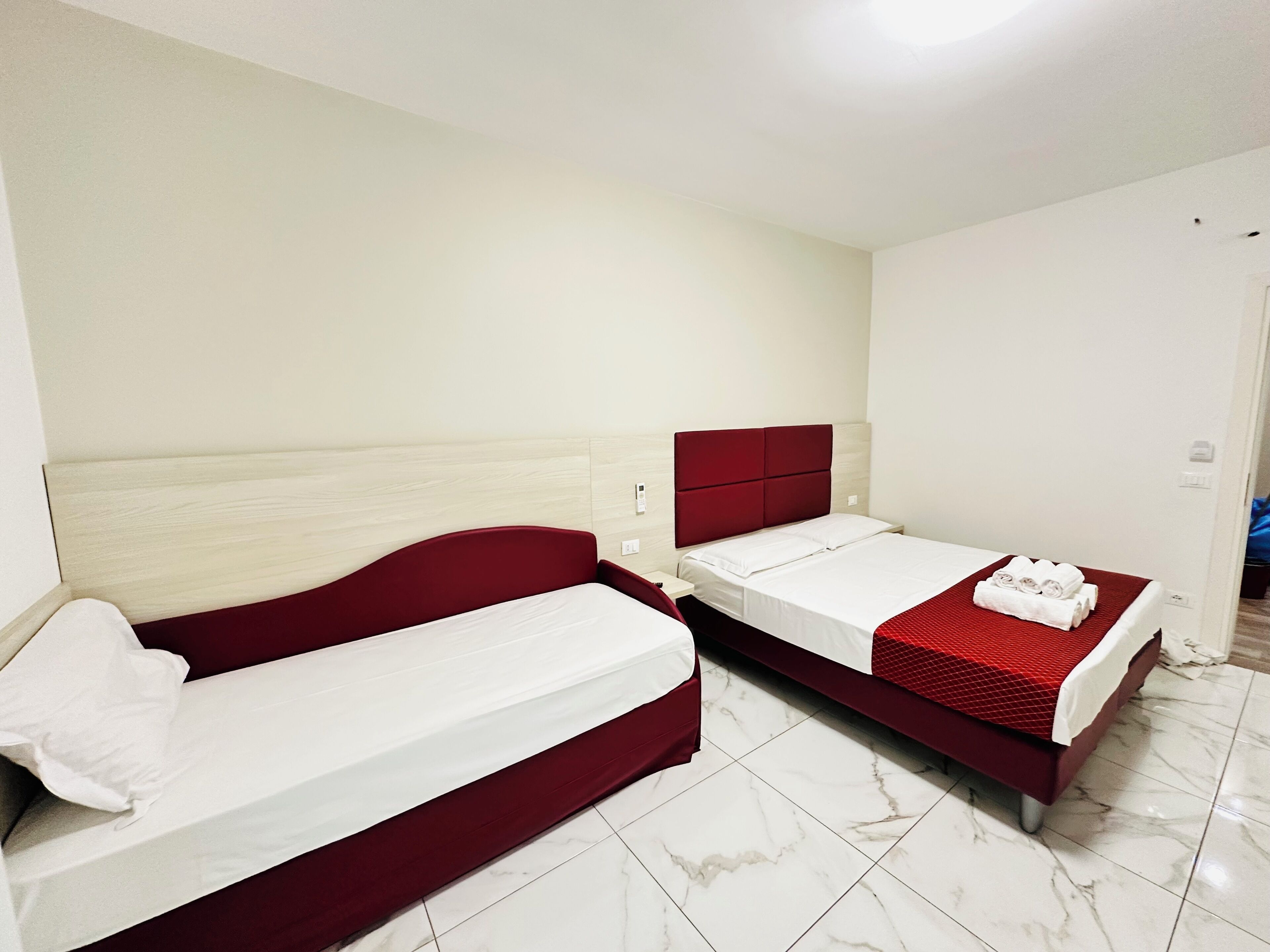 Ein Hotelzimmer mit zwei Einzelbetten, jeweils mit rotem Kopfteil und weißer Bettwäsche. Das Zimmer hat weiße Wände und einen gefliesten Boden.