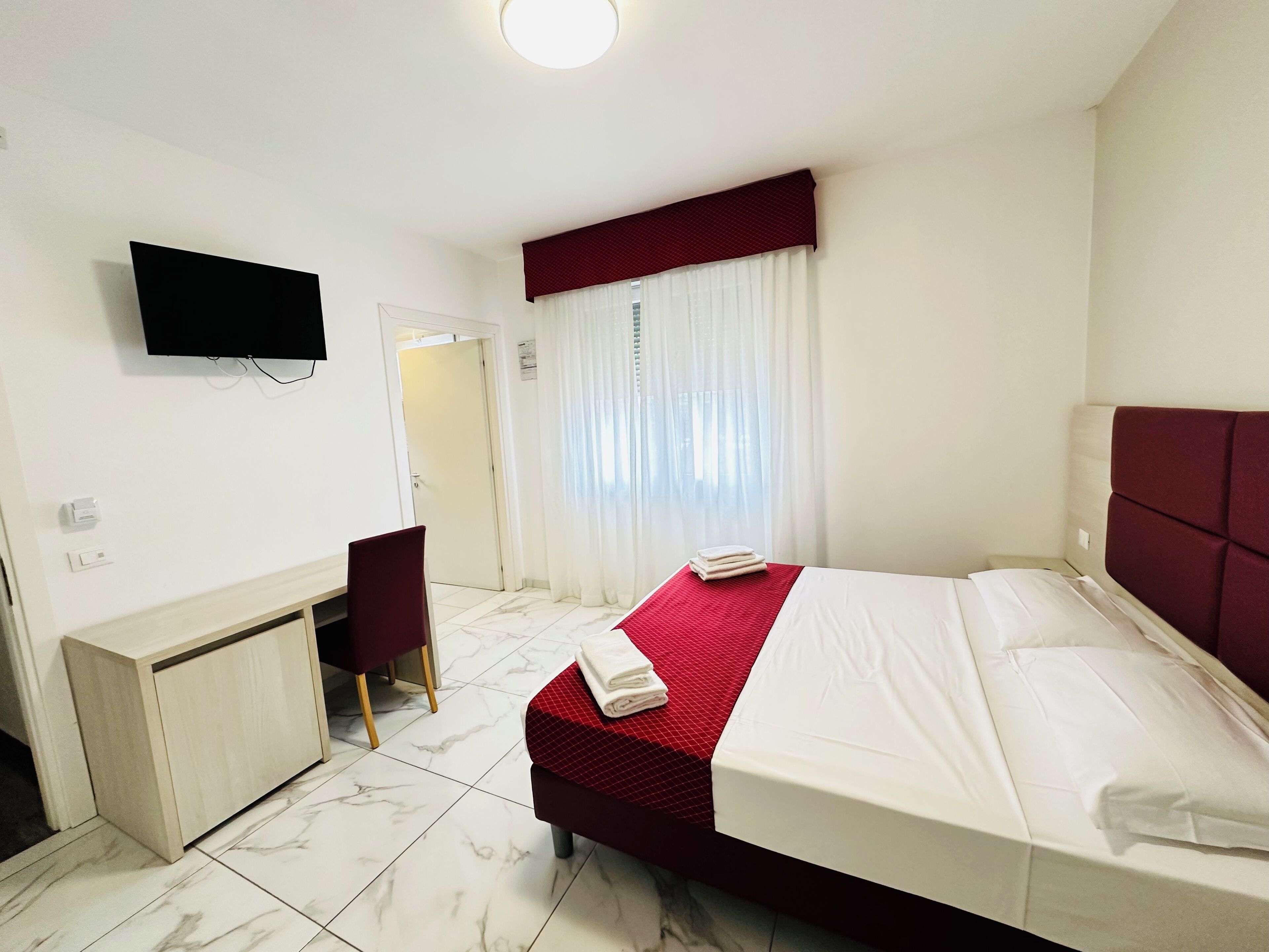 Ein Hotelzimmer mit Bett, Schreibtisch, Stuhl, Fernseher und einem Fenster mit Vorhängen.