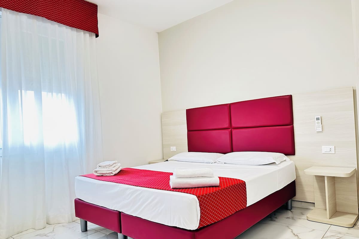 Une chambre d’hôtel avec un lit, une table de chevet, une tête de lit rouge et une fenêtre avec des rideaux blancs.