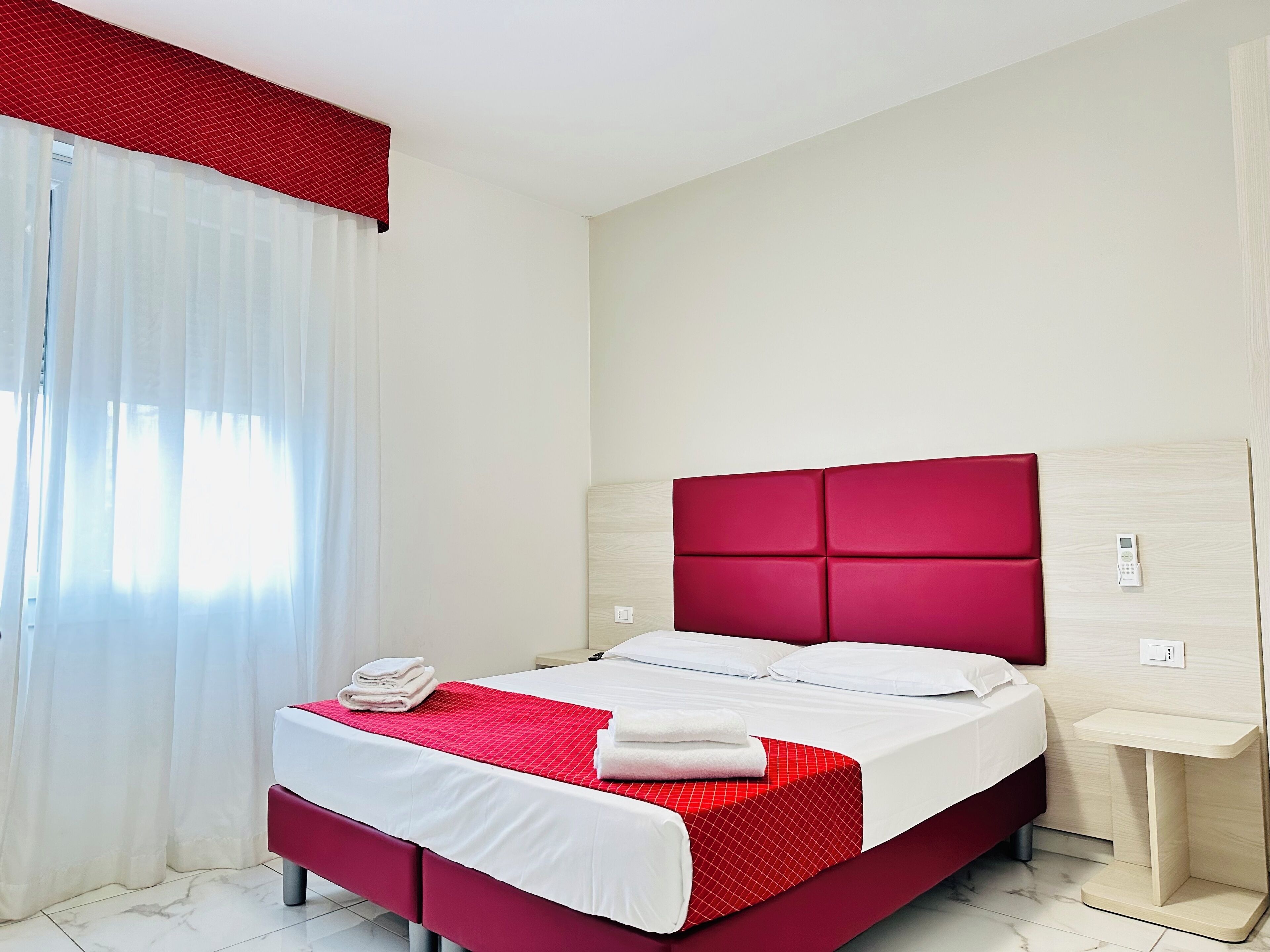 A hotel room with a bed, a bedside table, a red headboard, and a window with white curtains.
